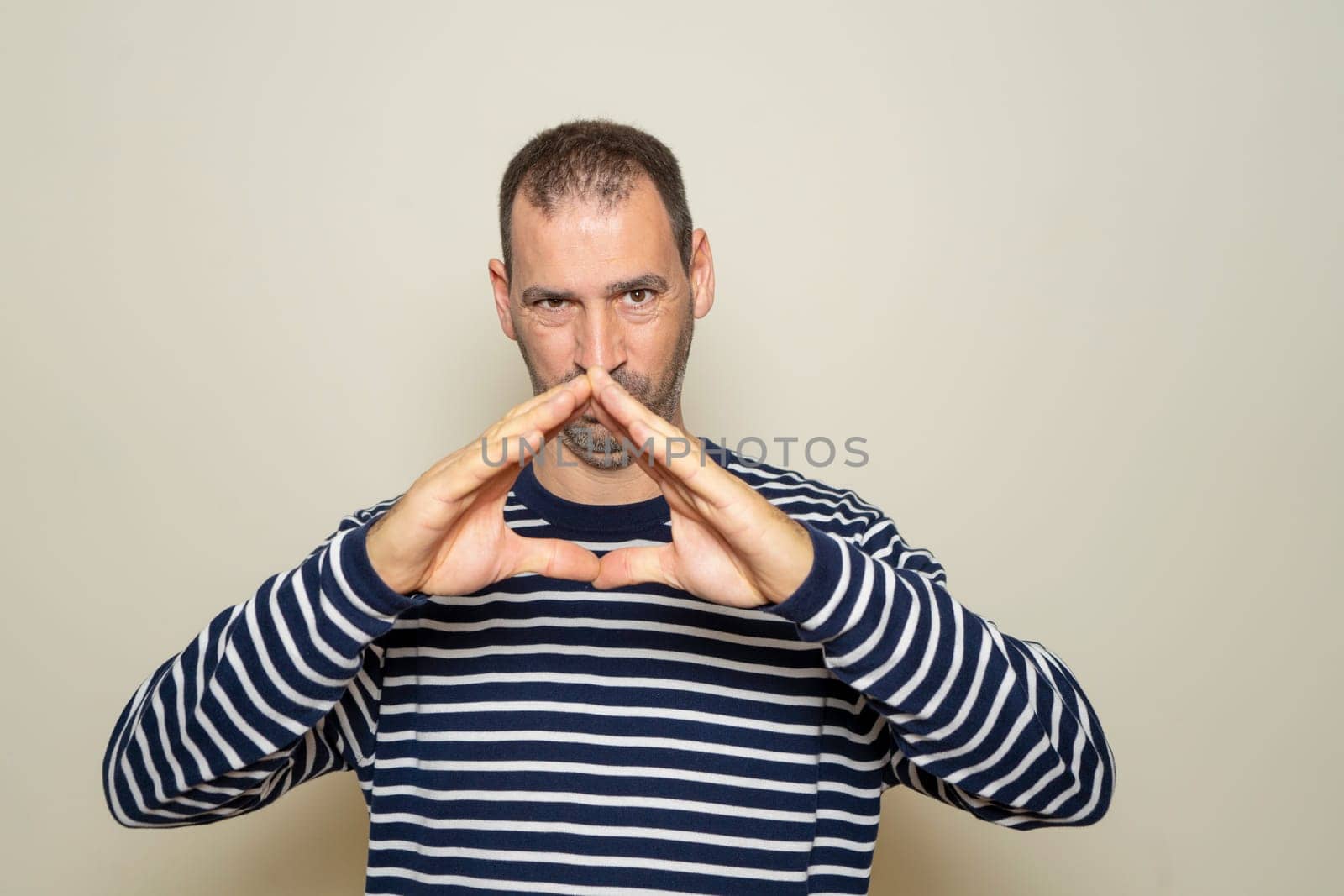 Hispanic man with a beard with his fingertips together in an expression of evil, cunning and cold planning a crime. Isolated on beige background. by Barriolo82