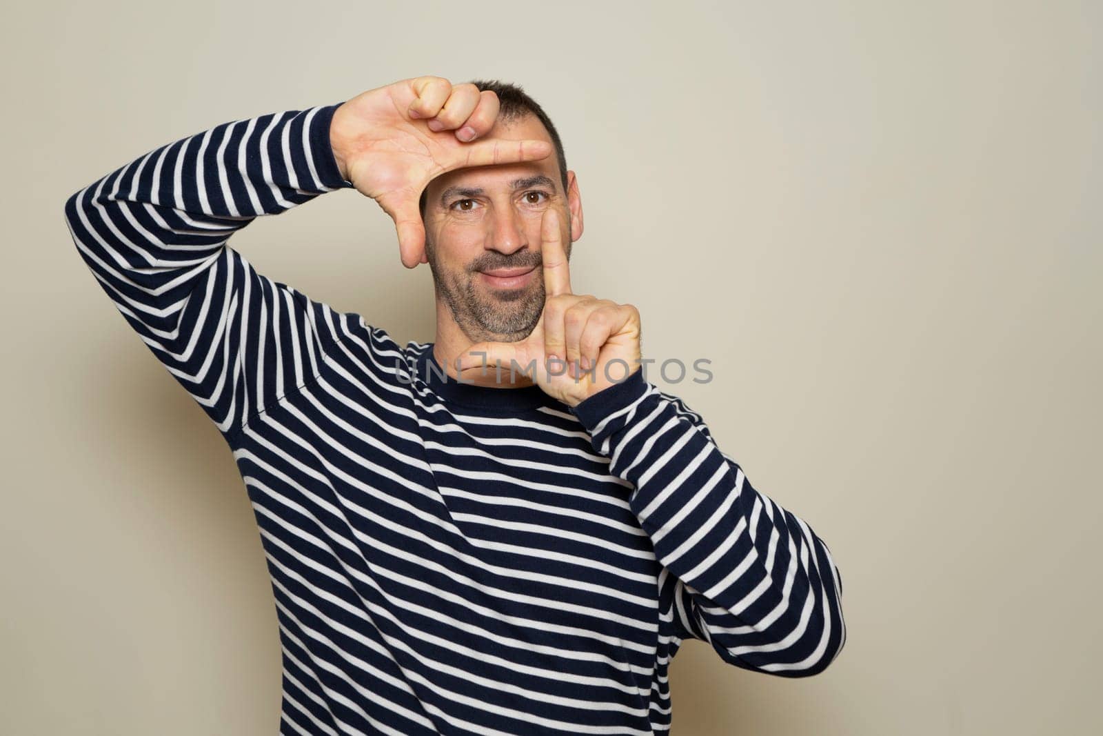A happy bearded man blinks his eyes and looks through a frame formed by his hands. He makes a frame with his fingers, isolated on beige background with copy space for text. People concept