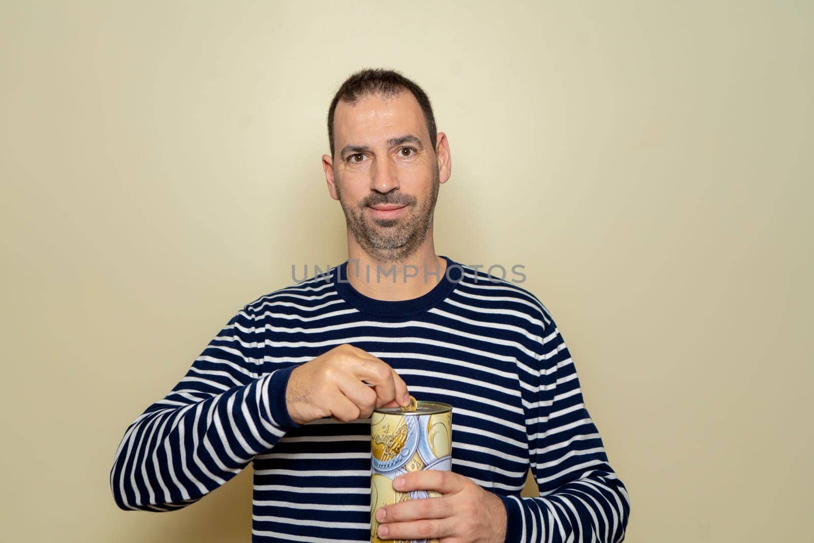 A Hispanic man with a beard wearing a striped sweater putting a euro coin into a metal piggy bank, trying to save so he can travel on vacation. Isolated on beige studio background. by Barriolo82