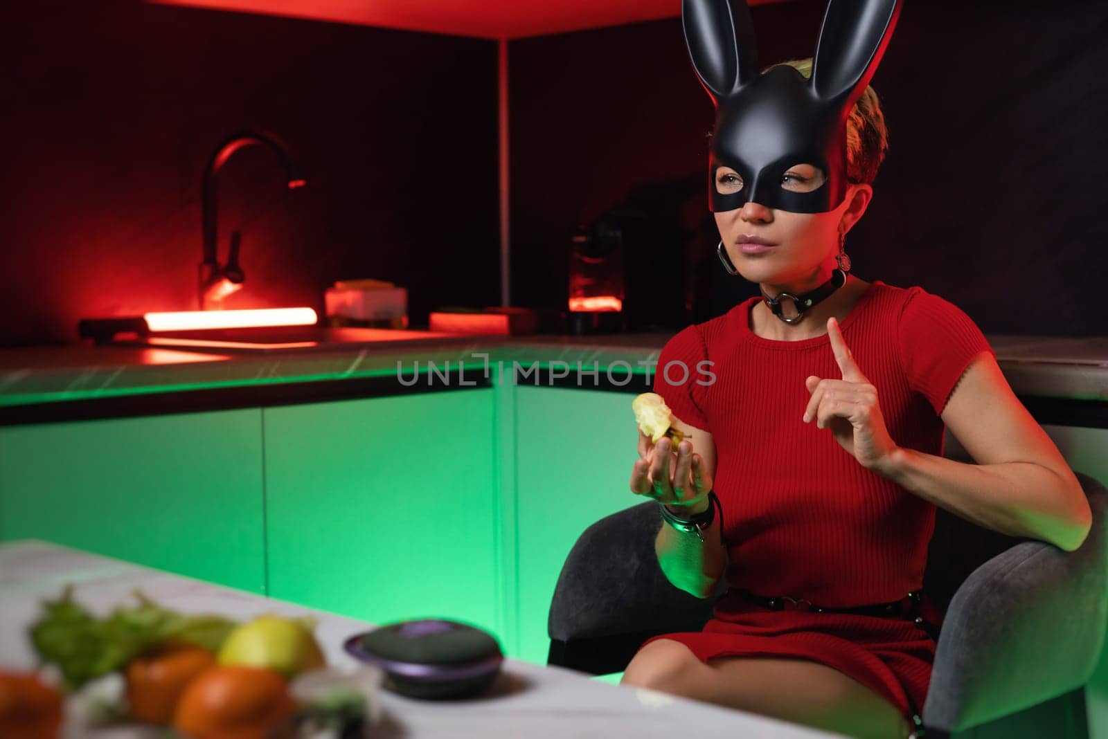 beautiful girl in a bdsm rabbit mask and a bright red dress eats an apple in the kitchen in neon light promoting a healthy lifestyle vegetarianism by Rotozey