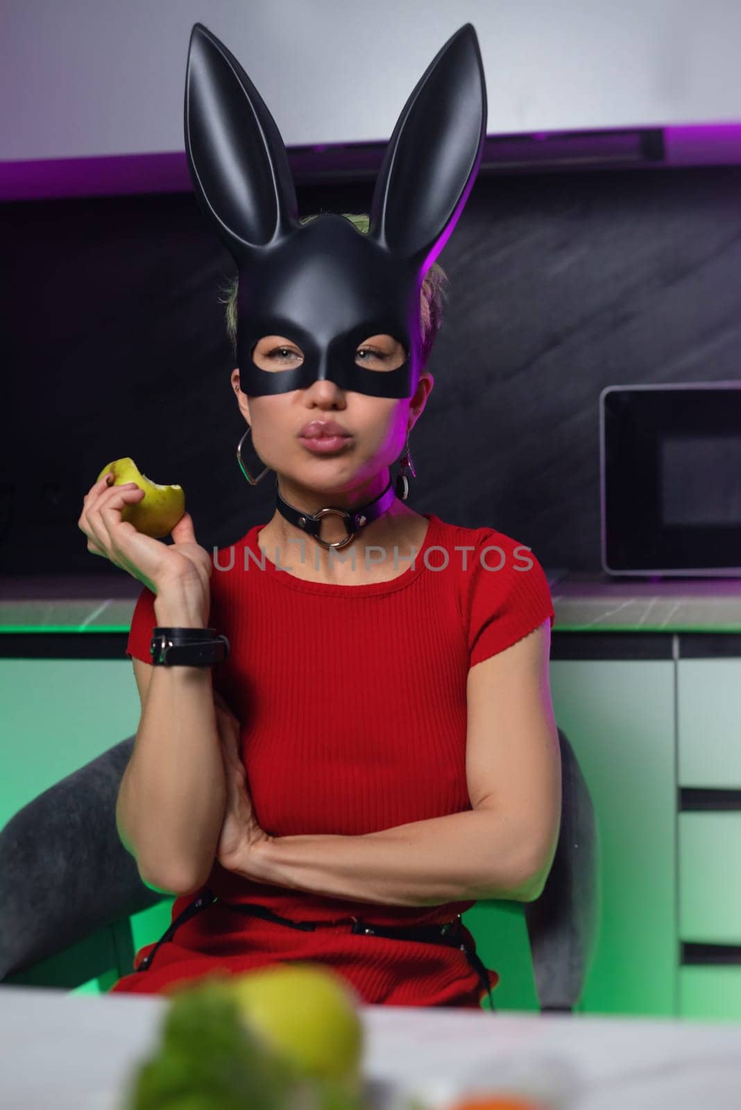 beautiful girl in bdsm rabbit mask and a bright red dress eats an apple in the kitchen in neon light promoting a healthy lifestyle vegetarianism