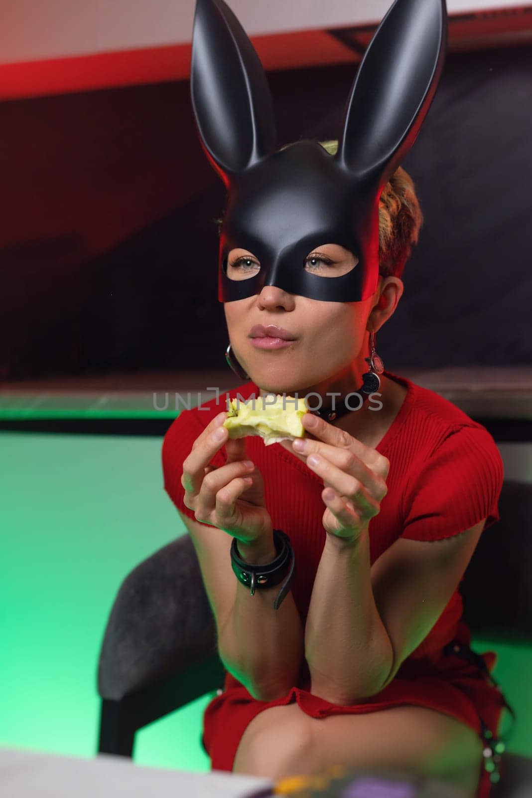 beautiful girl in a bdsm rabbit mask and a bright red dress eats an apple in the kitchen in neon light promoting a healthy lifestyle vegetarianism by Rotozey