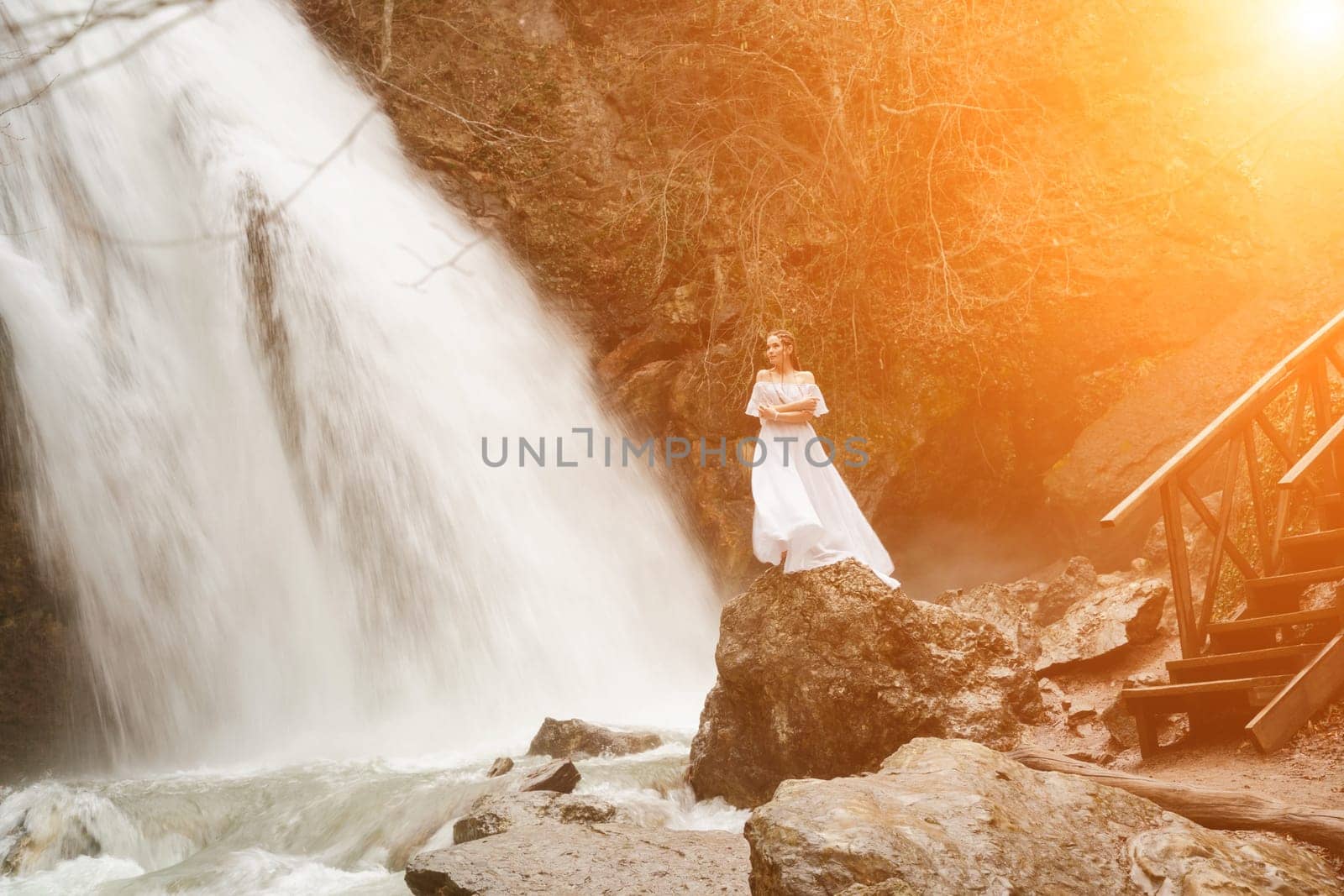 Happy woman in a white dress stands on a stone with a waterfall behind by Matiunina