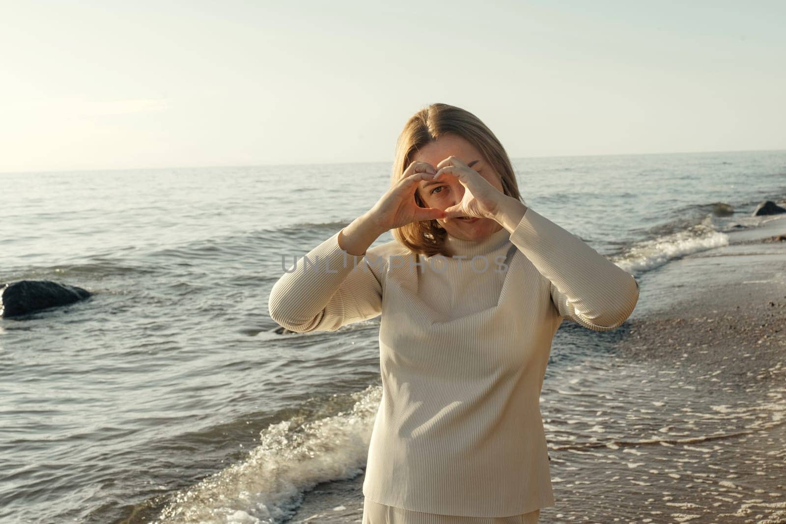 A woman stands on the shore, playfully depicting a heart with her hands, against the backdrop of gentle waves and a clear sky.