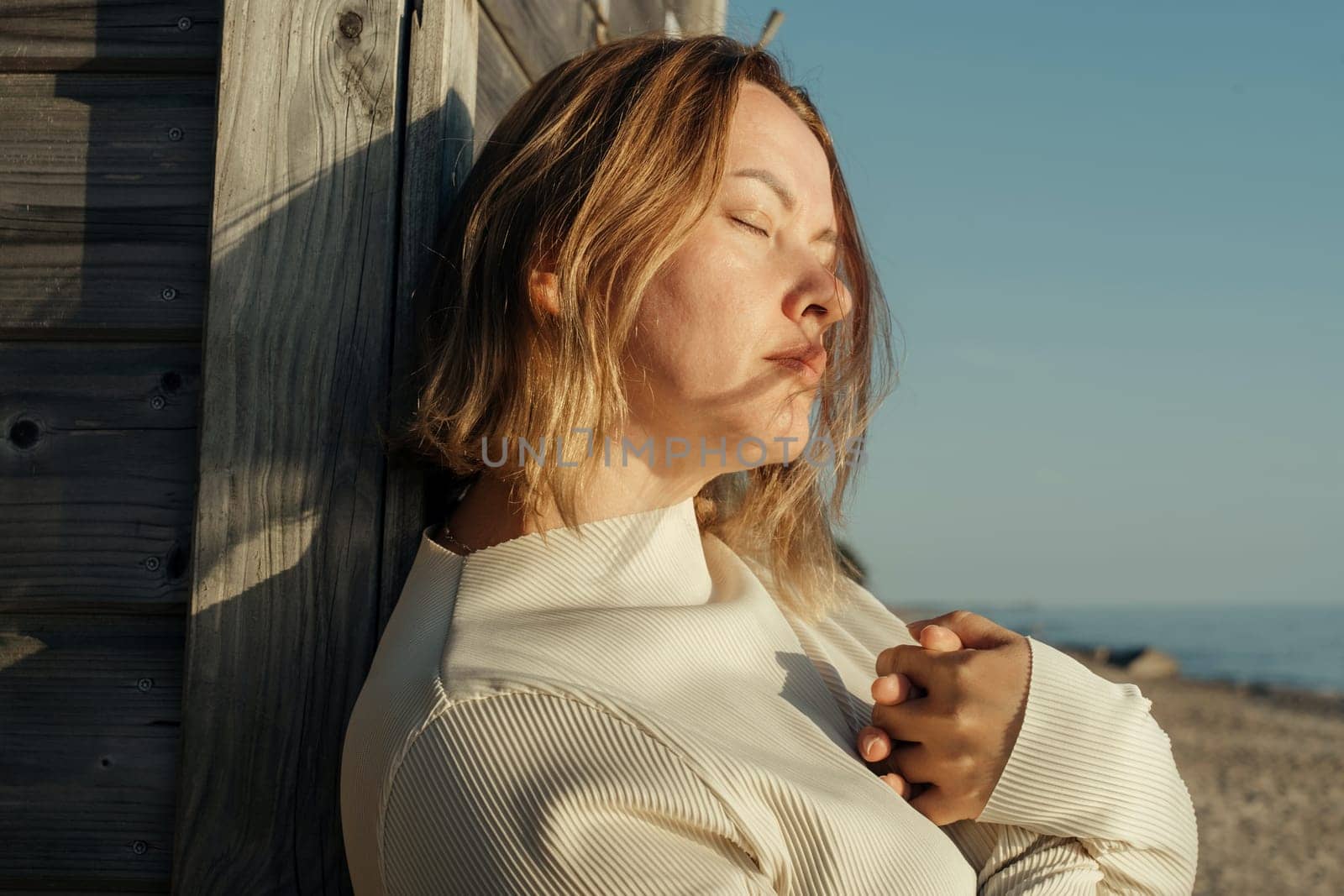 A woman leans casually against a weathered wooden wall near the ocean, gazing out at the waves crashing on the shore.
