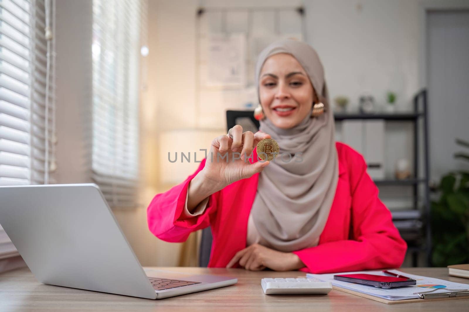 A female Muslim investor who trades in cryptocurrencies sits looking at stock charts and studying Bitcoin..