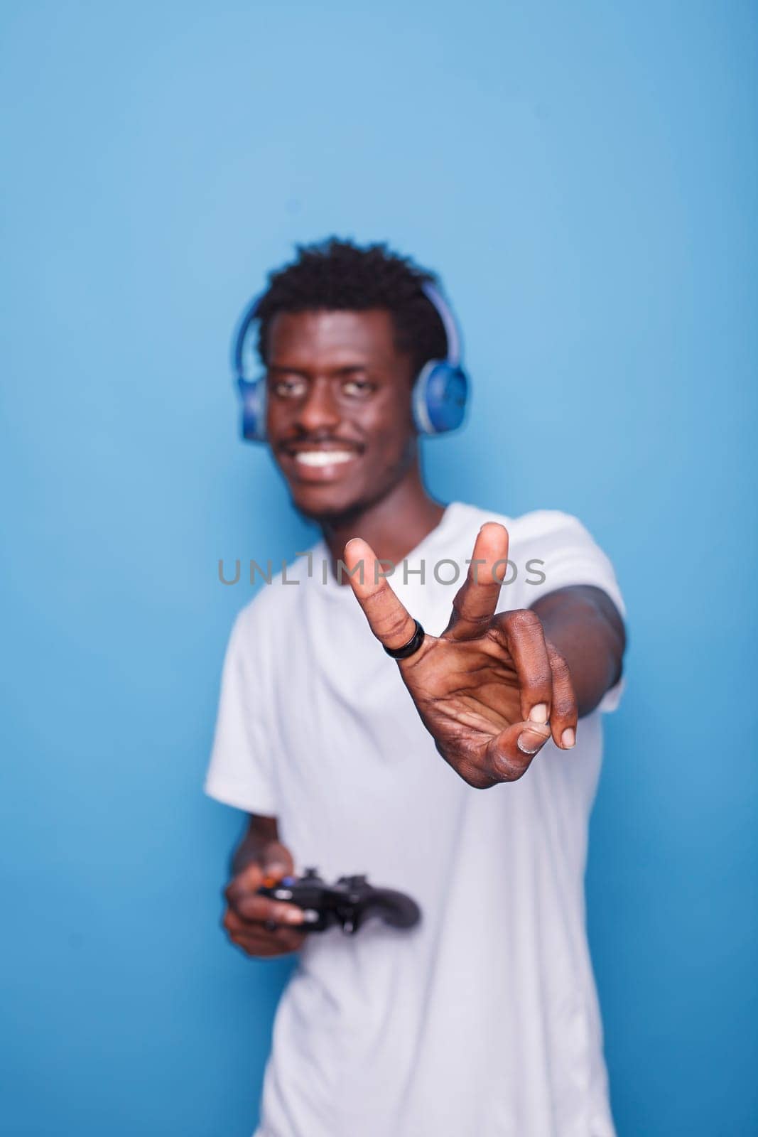 African American man wearing headphones and carrying controller gesture peace sign towards camera. Black person standing in front of isolated backdrop makes happy gesture with their hand.