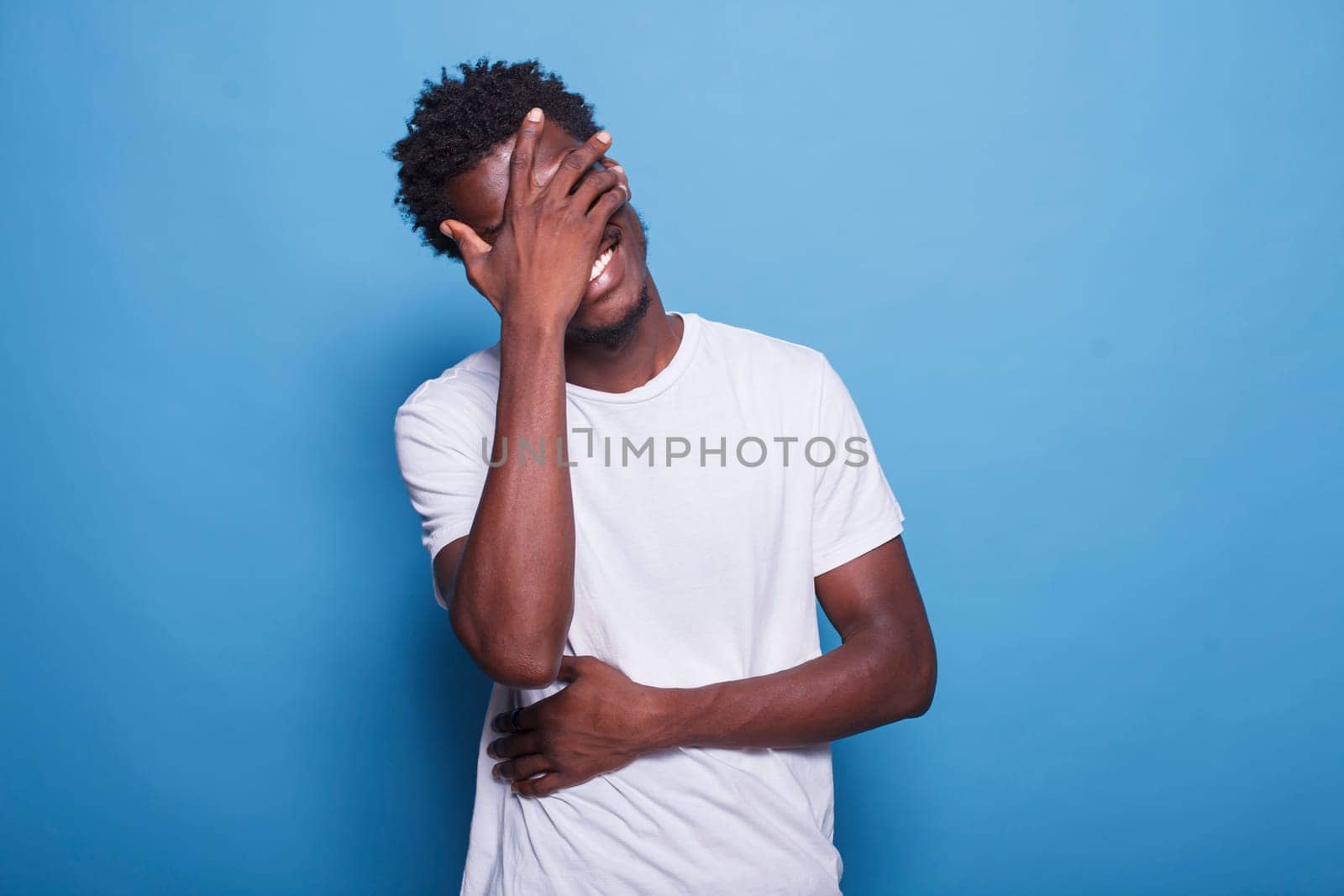 Portrait of joyful young man with afro, laughing at camera while standing in studio with blue background. African american guy with his hands covering eyes and stomach is smiling and chuckling.