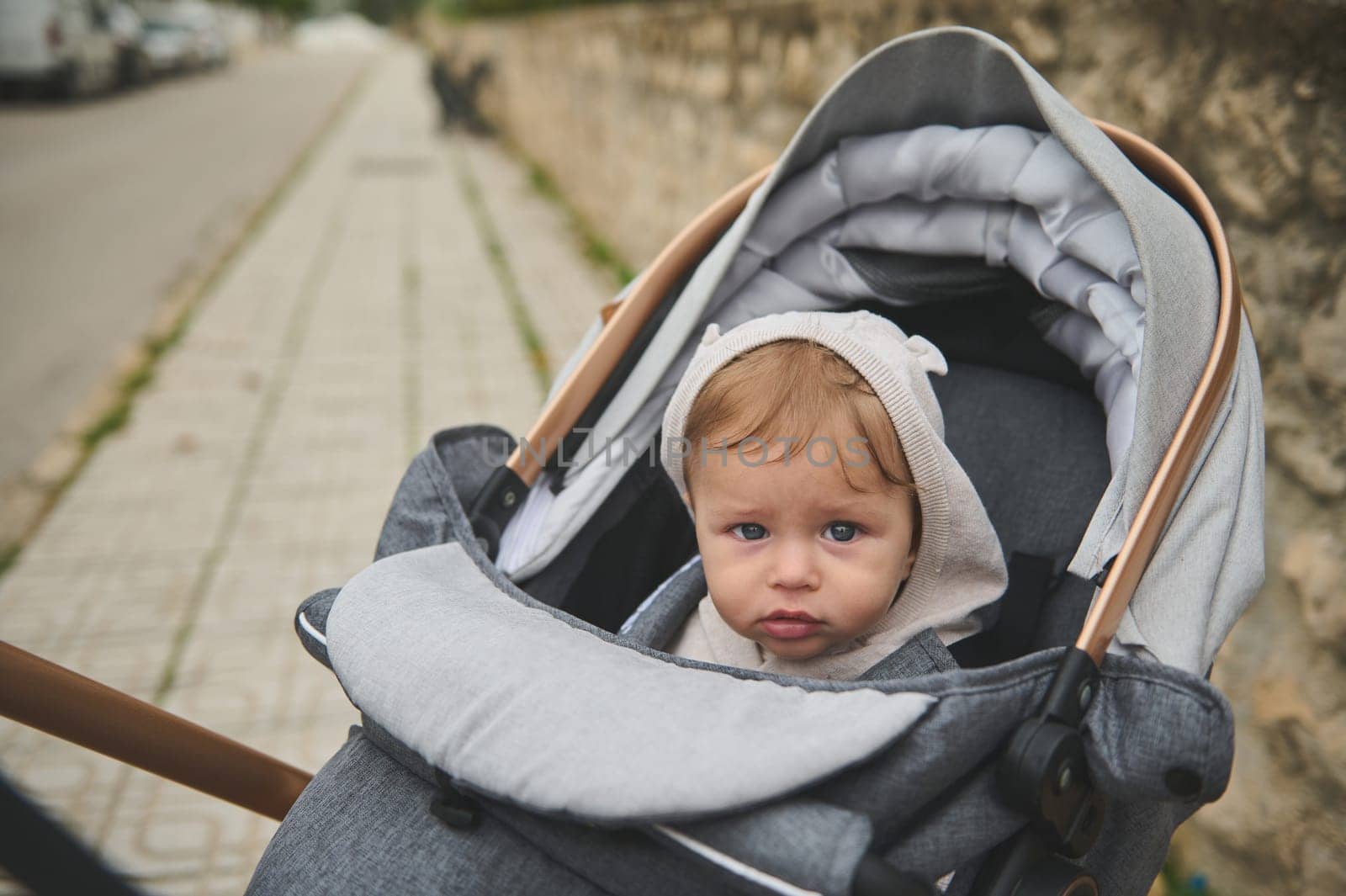 Authentic portrait of an European adorable baby boy 7-9 months old, with hood on his head, looking at the camera, sitting in a baby stroller outdoors