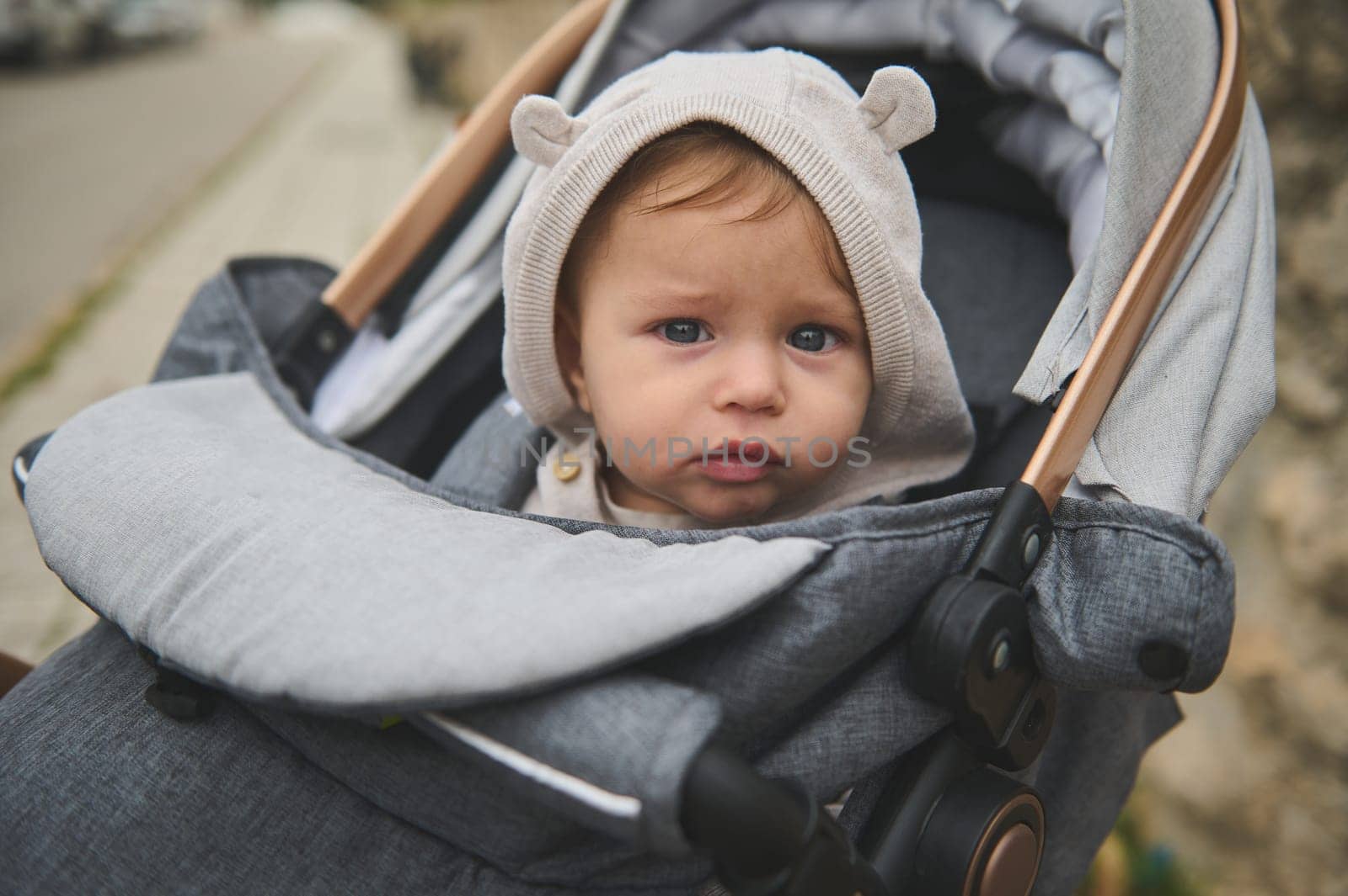 Portrait of a Caucasian adorable baby boy with hood on his head, looking at the camera, sitting in a baby pram outdoors