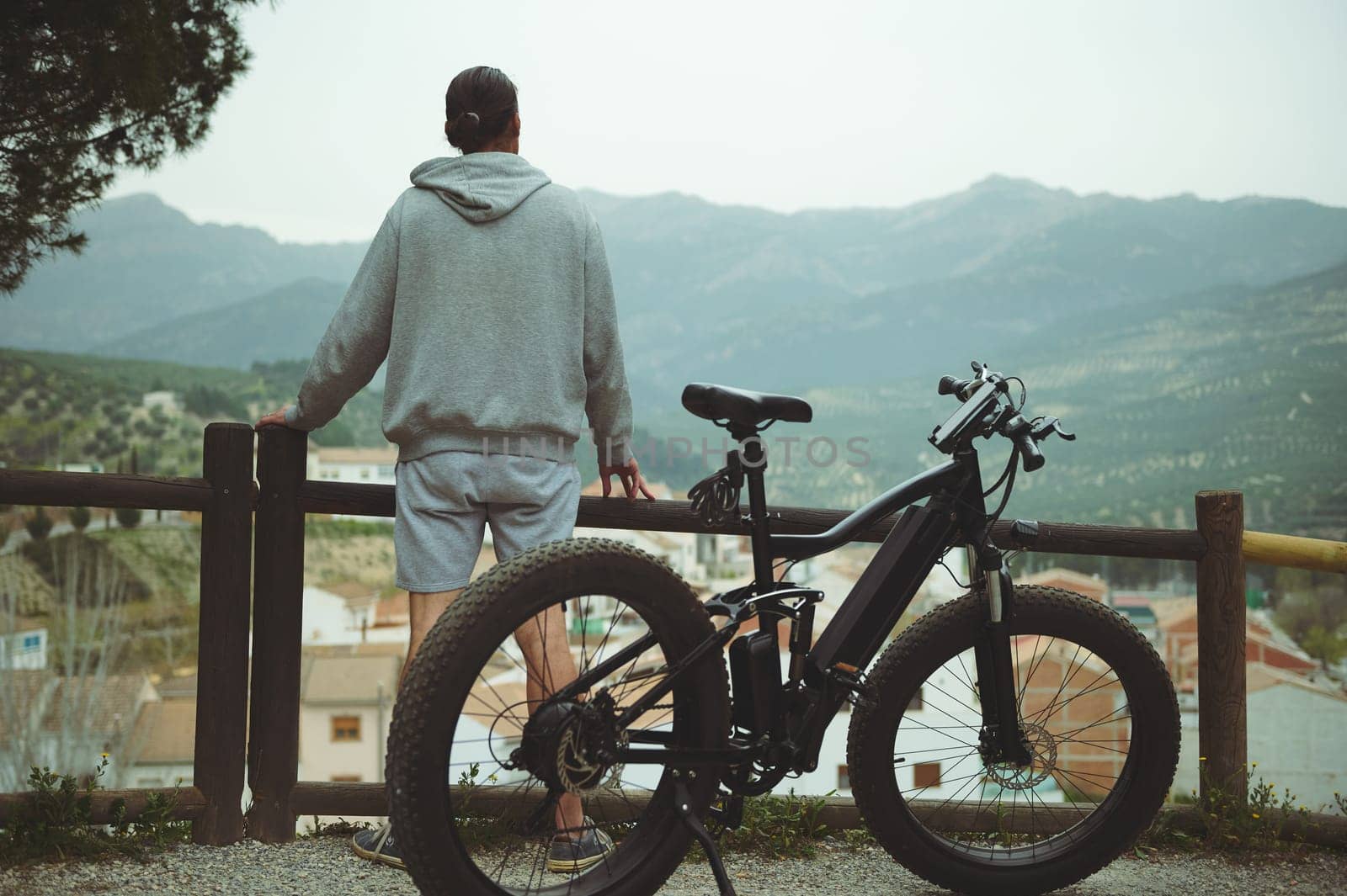 Healthy lifestyle. Sport. Inspiration. Achievements. Rear view young active man with electric bicycle standing on top of hill and contemplating a beautiful view of a city in the mountains on foggy day