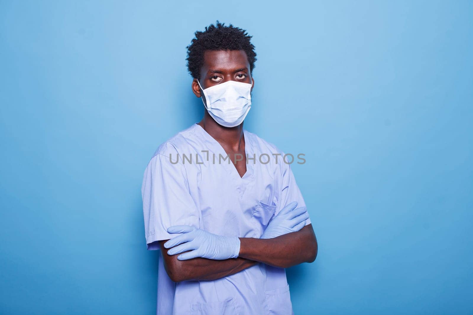 In studio, nurse with crossed arms, wearing face mask and gloves, looks at camera. Black man working as medical assistant stands over isolated backdrop, protected against covid 19 pandemic.