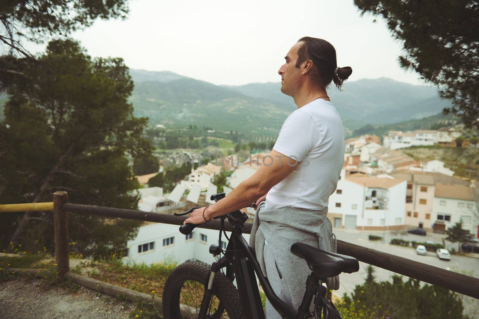 Side view of a male cyclist in white t-shirt, having a break, standing by his electric bike, admiring the medieval village from the top of valley by artgf