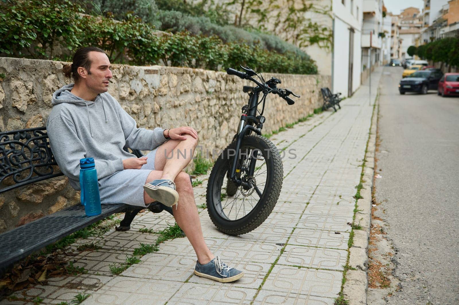 Young Caucasian happy man cyclist sitting on a bench near his electric bike, relaxing after ride in mountain city, enjoy an active weekend outdoors, wearing gray sports clothes. Copy advertising space