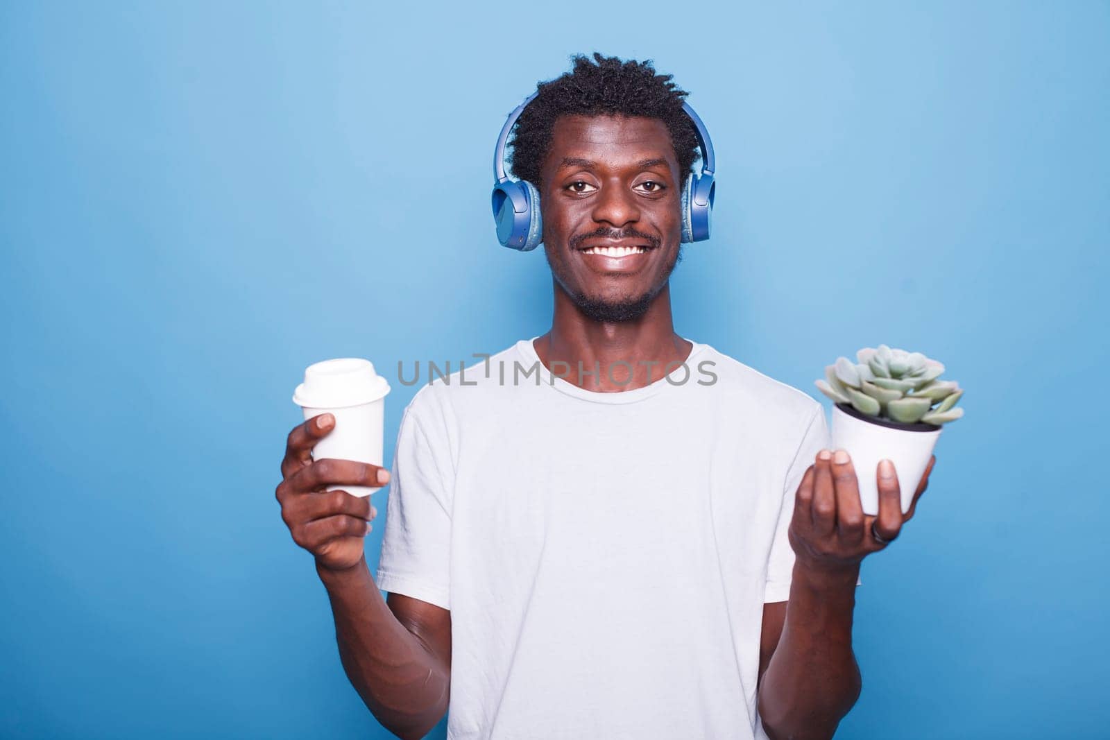 Portrait of happy African American man standing in front of a blue background with a potted plant while carrying a beverage. Smiling black guy holding a coffee cup and wearing wireless headphones.
