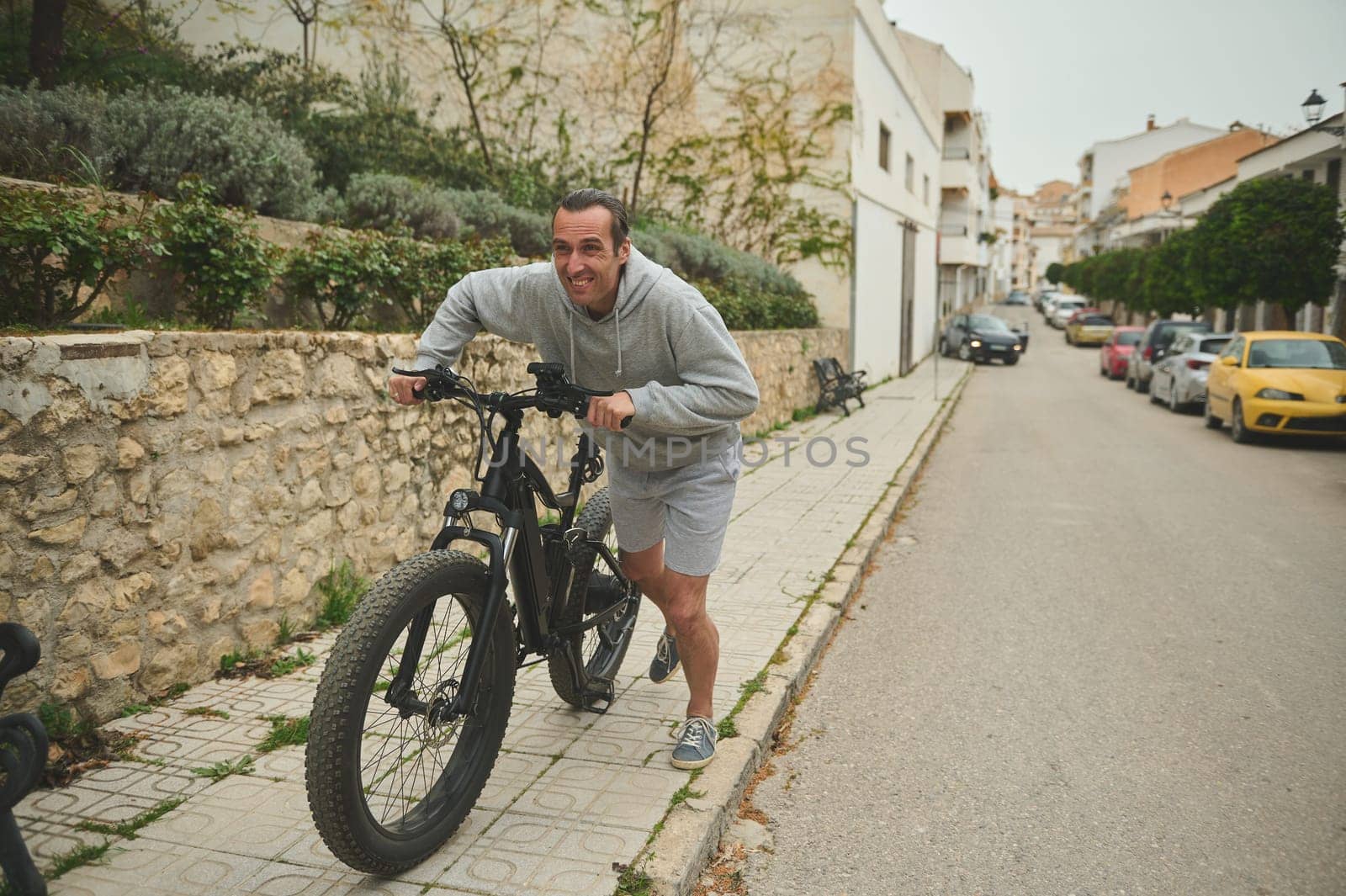 Caucasian young man 40 years old, pushing electric mountain bike up the street. Active lifestyle. Sport. Adventure travel on bicycle. Using el-bicycle ad ecologically friendly mode of urban transport