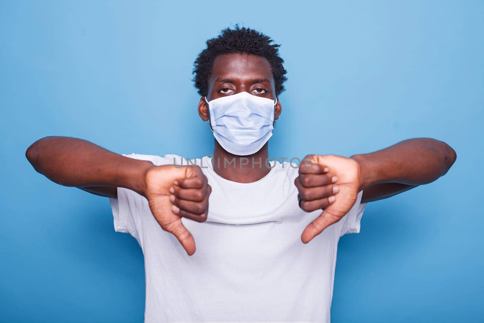 African American adult showing thumbs down sign with both hands. Black man gesturing with his fingers while looking at camera and wearing face mask to protect from coronavirus epidemic.