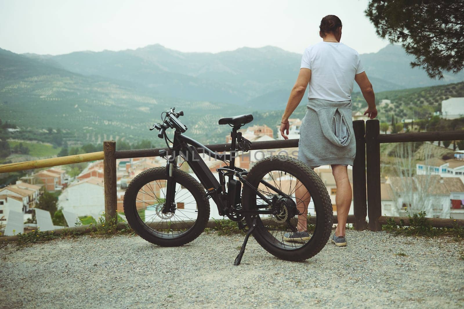 Rear view of a young man cyclist, standing near his electric bike, having a break after biking in mountains. People. Active healthy lifestyle. E-bike as sustainable, eco friendly transport