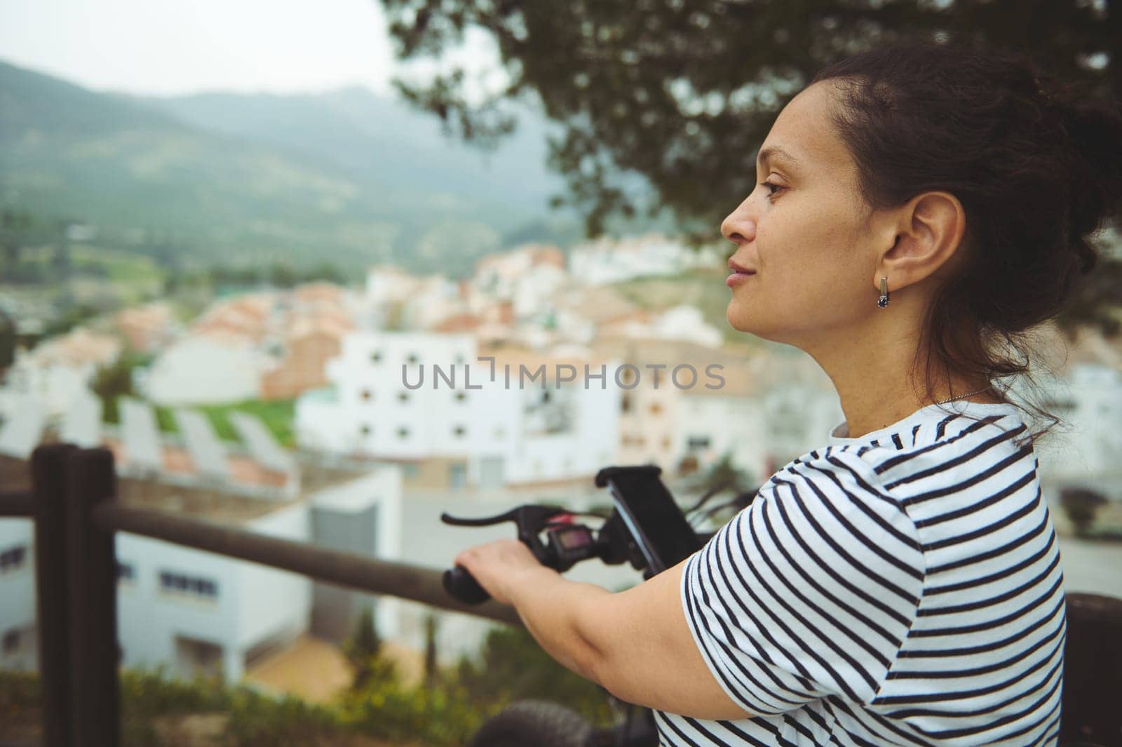 Close-up side portrait of a young multi ethnic woman cyclist in white and black striped t-shirt, having a break, standing by her electric bike, admiring the medieval village from the top of valley by artgf