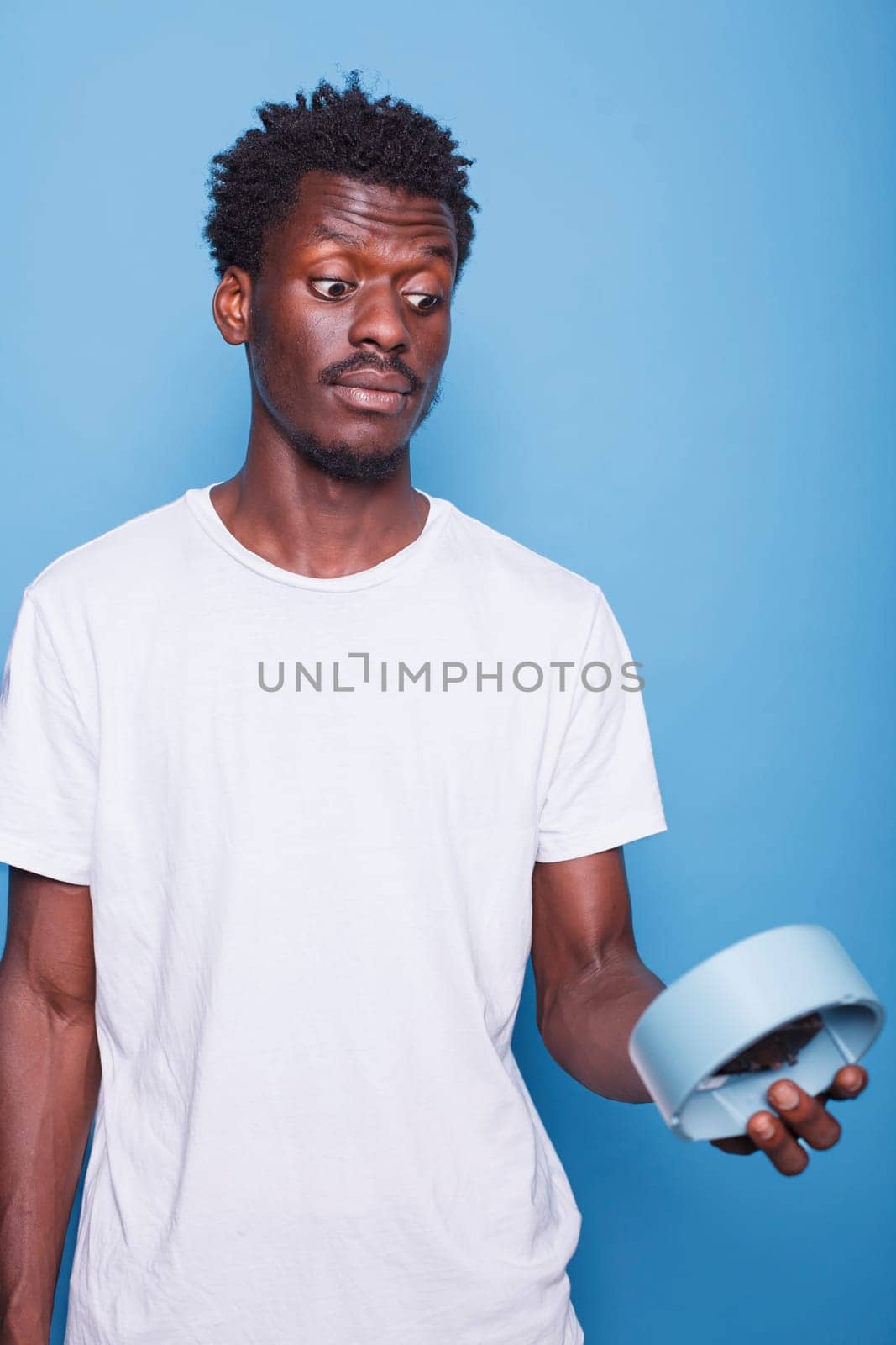 Close-up of african american man carrying wall clock with shocked expression. Portrait of black male individual with surprised face, looking at hours and minutes on a blue watch for alarm.