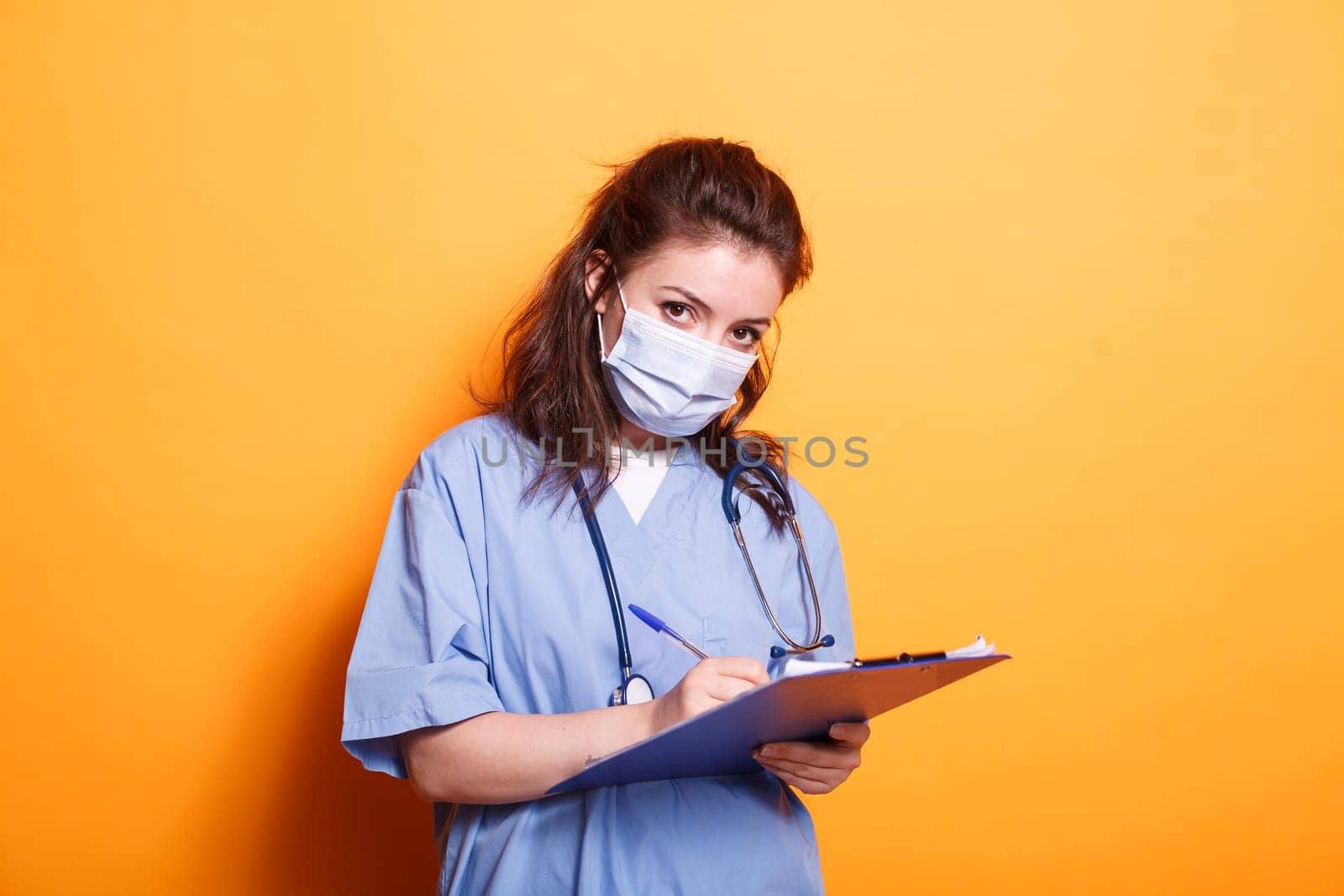 Woman doctor in blue scrubs taking notes on clipboard files, standing against isolated background. Healthcare specialist with face mask writing on medical documents, looking at camera.