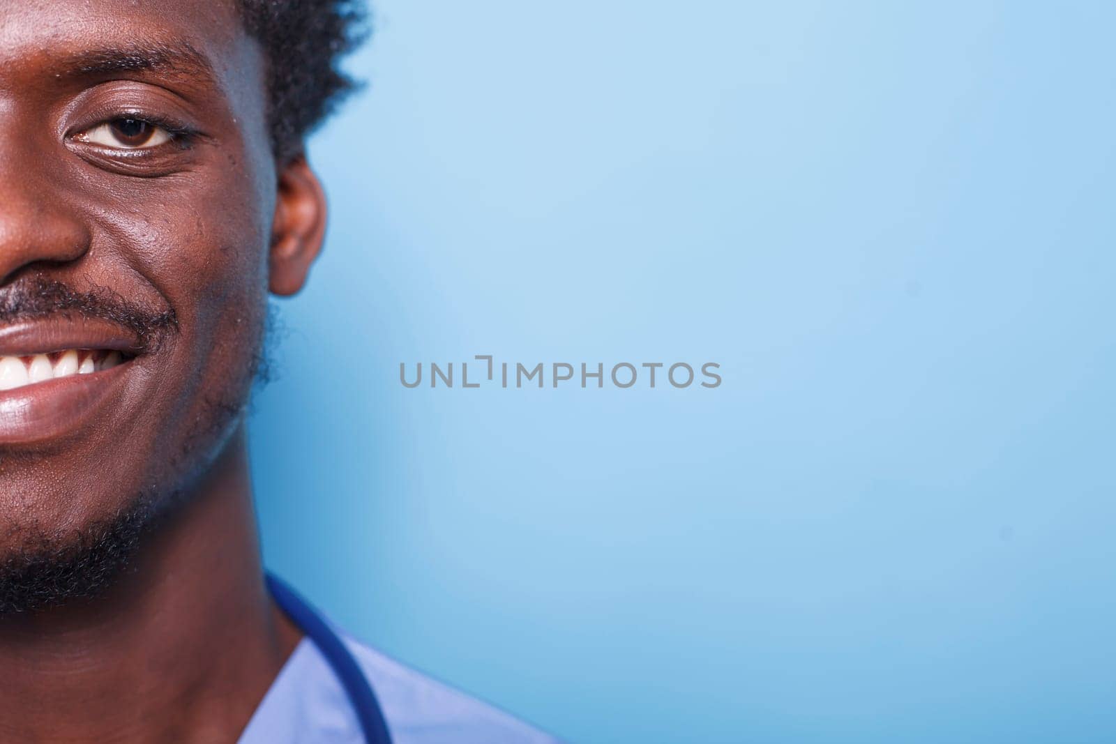 Close-up portrait of cheerful male nurse wearing scrubs and stethoscope on a blue background. Half face, cropped image of confident African American medical physician smiling at camera.