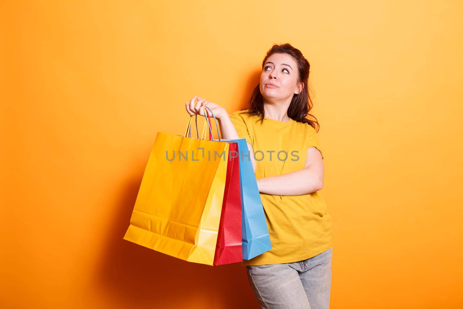 Happy, stylish brunette shopper carries shopping bags and smiles on isolated orange background. Caucasian woman looking attractive and cheerful while grasping colorful parcels.