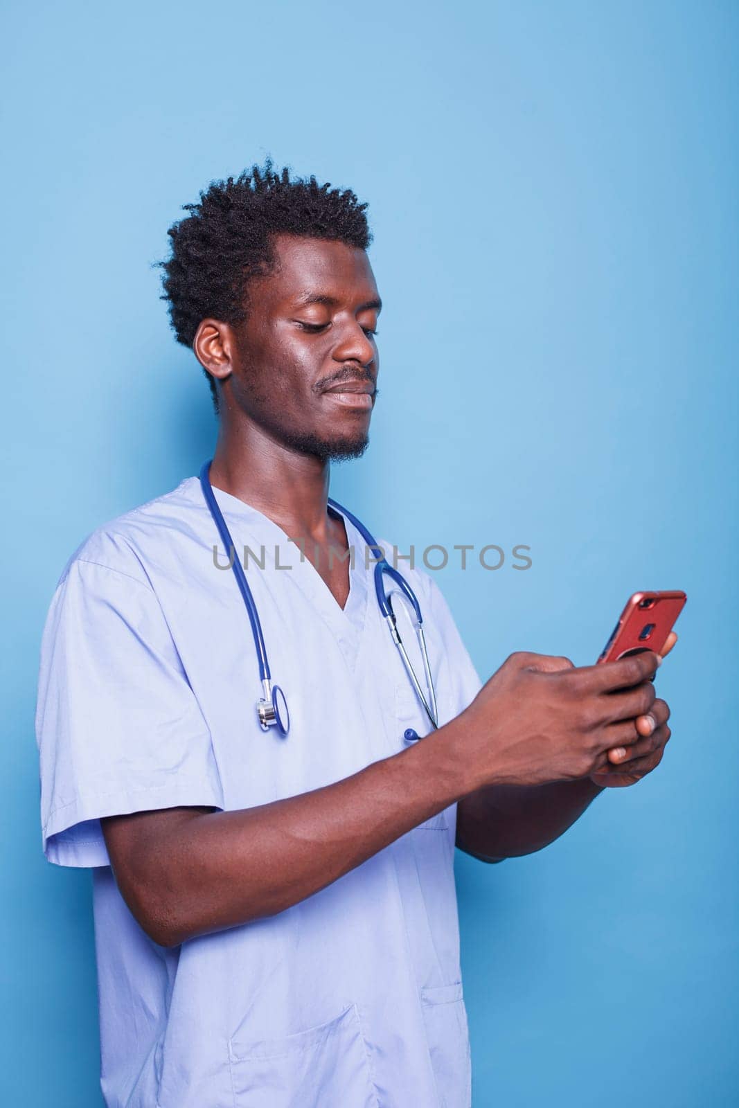 African American doctor in scrubs wears stethoscope and holds a smartphone, smiling confidently in a studio. Black modern healthcare professional utilizing mobile device during covid 19 pandemic.