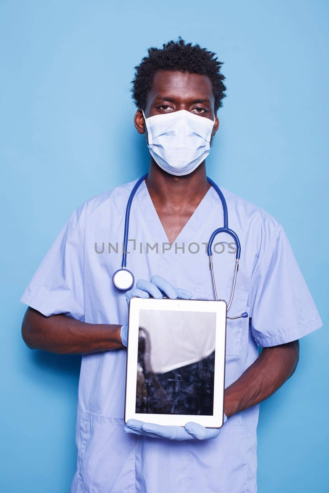 Male nurse looking at camera while holding a tablet with blank screen, against blue background. Demonstrating healthcare expertise, black man wears scrubs, face mask, gloves, and stethoscope.