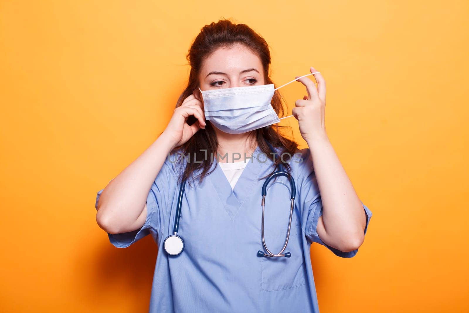 Nurse practitioner putting on face mask for protection in front of camera. Female medical doctor wearing scrubs and stethoscope having safety mask against coronavirus during pandemic.