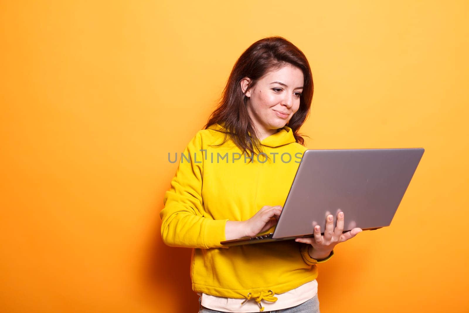 In the studio, young adult looks at a laptop to use technology on camera. Portrait of a modern woman standing over an isolated background, holding a digital device and surfing the internet.