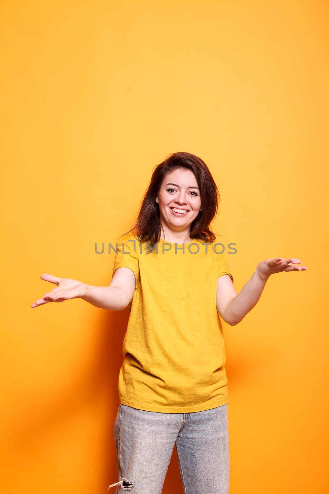 Joyful young woman with expressive positive attitude welcoming with raised arms in studio. Portrait of brunette lady smiling, being friendly and sociable, doing a greeting gesture.