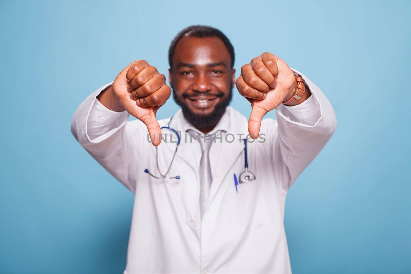 Black man wearing a hospital uniform and stethoscope standing and doing thumbs down to the camera. Portrait of african american medical specialist making disapproval hand gestures.