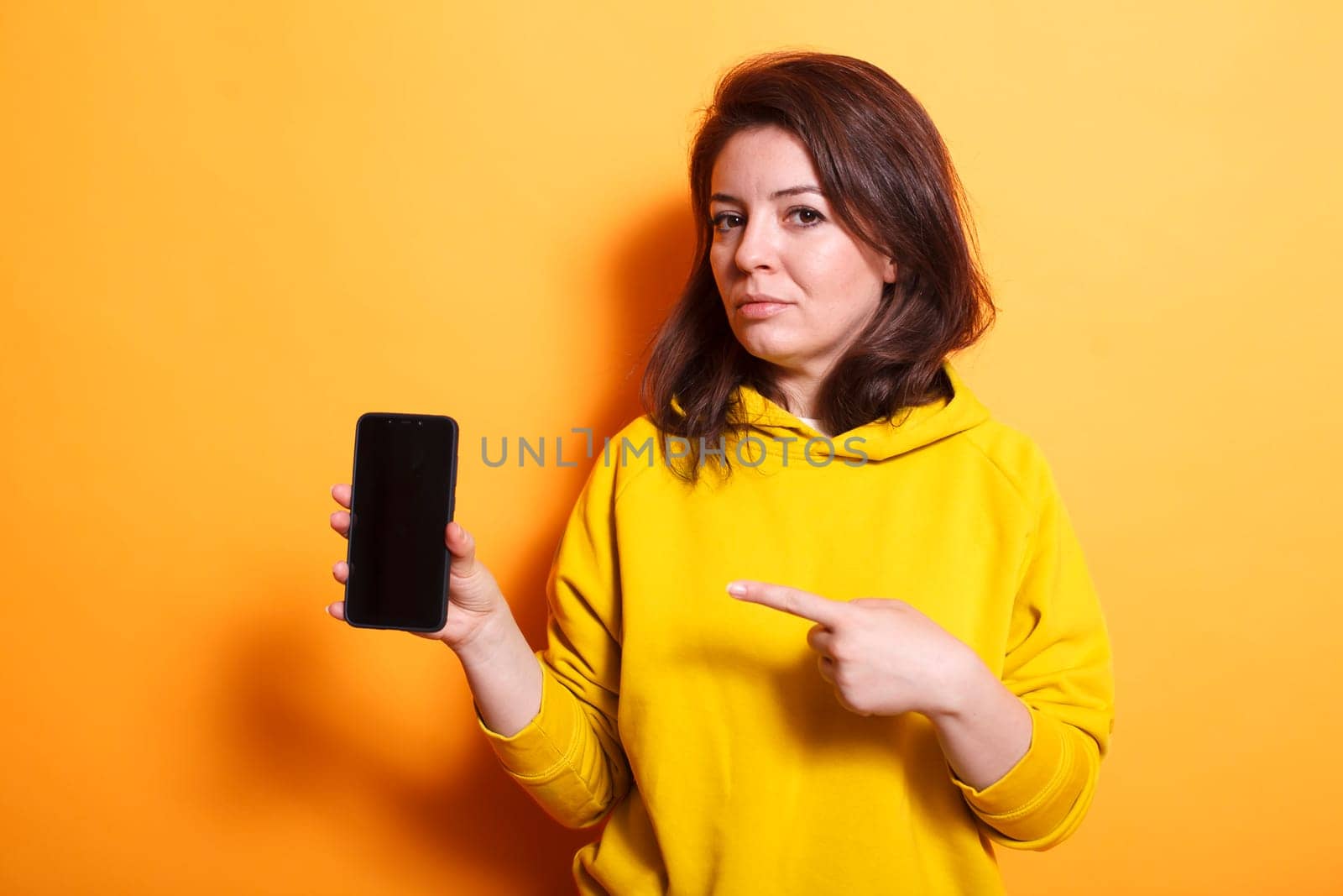 Brunette lady showing cellphone with empty screen at camera in studio. Woman pointing at mobile phone with isolated mockup template on display. Person standing over orange background