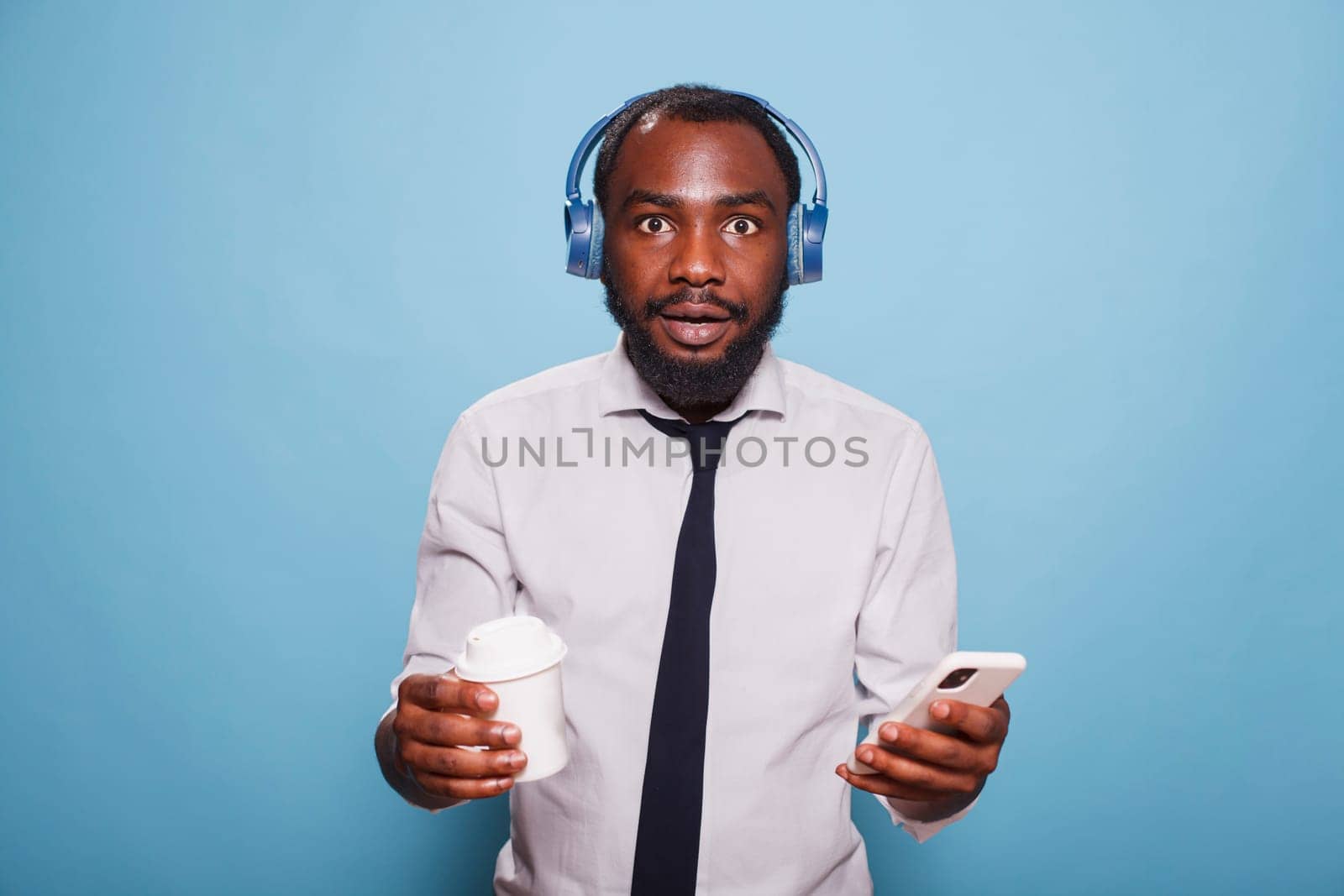 Shocked black man in white shirt looking at camera while holding cell phone for streaming music to wireless headphones. Young freelancer with a takeaway cup, having coffee and browsing internet.