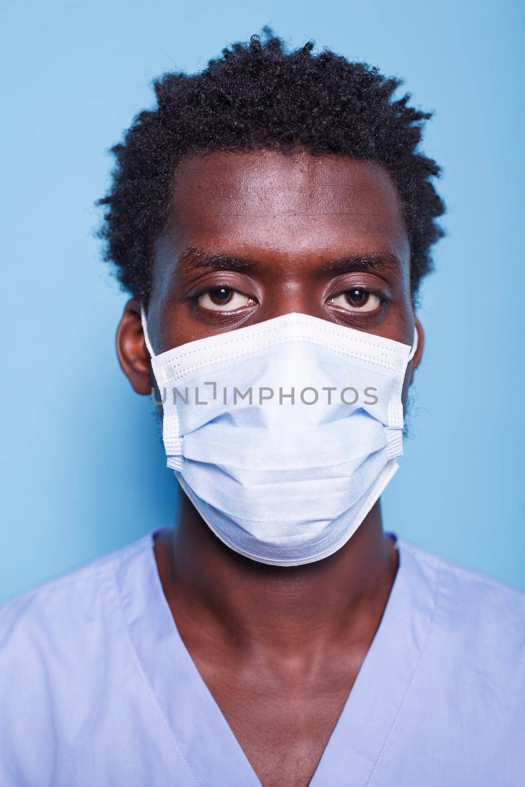 Close-up of medical assistant with face mask looking at camera in a studio. Portrait of African American man working as nurse, wearing blue uniform and having protection against covid 19.