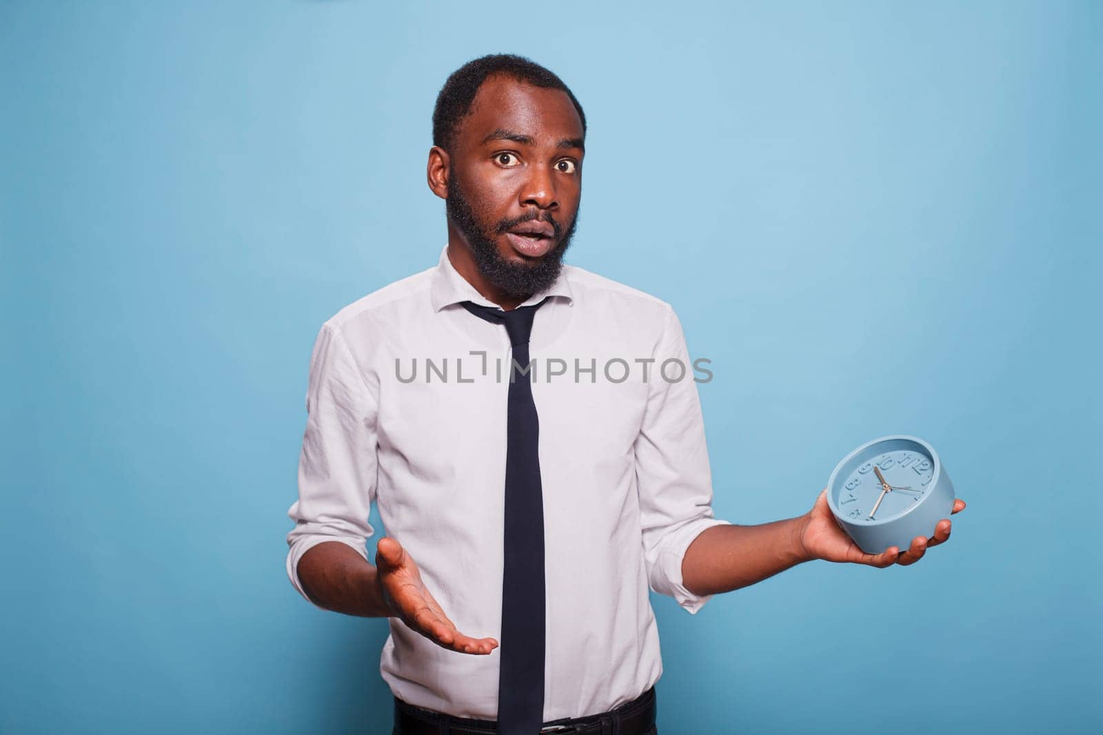 Young businessman looking surprised running out of time holding alarm clock while wearing white shirt and black tie. African american office worker panicking for being late for a meeting.