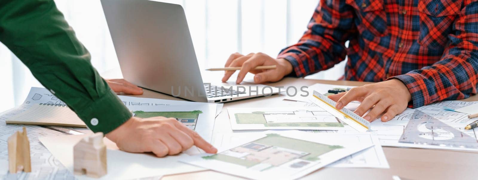 Professional architect design a blueprint by using laptop during project manager shows mistake point on table with architectural document and wooden block scatter around.. Closeup. Delineation.