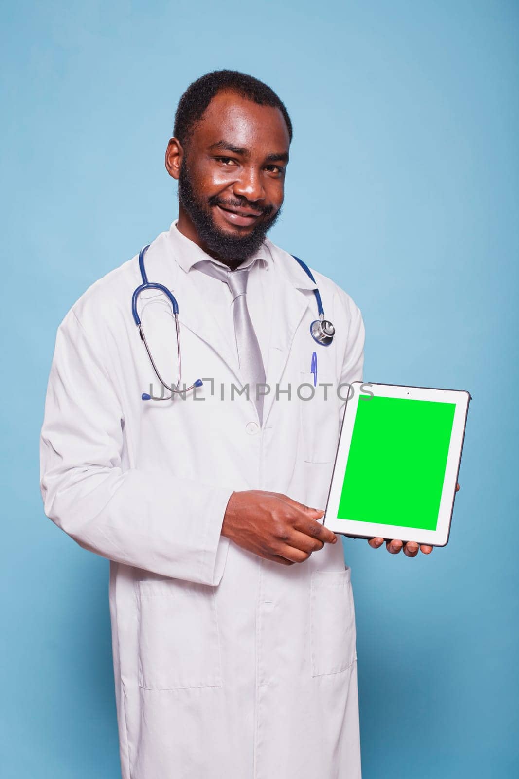 Portrait of black man wearing a stethoscope and lab coat is holding a tablet with green screen. African american doctor grasping device displaying blank chromakey mockup template.