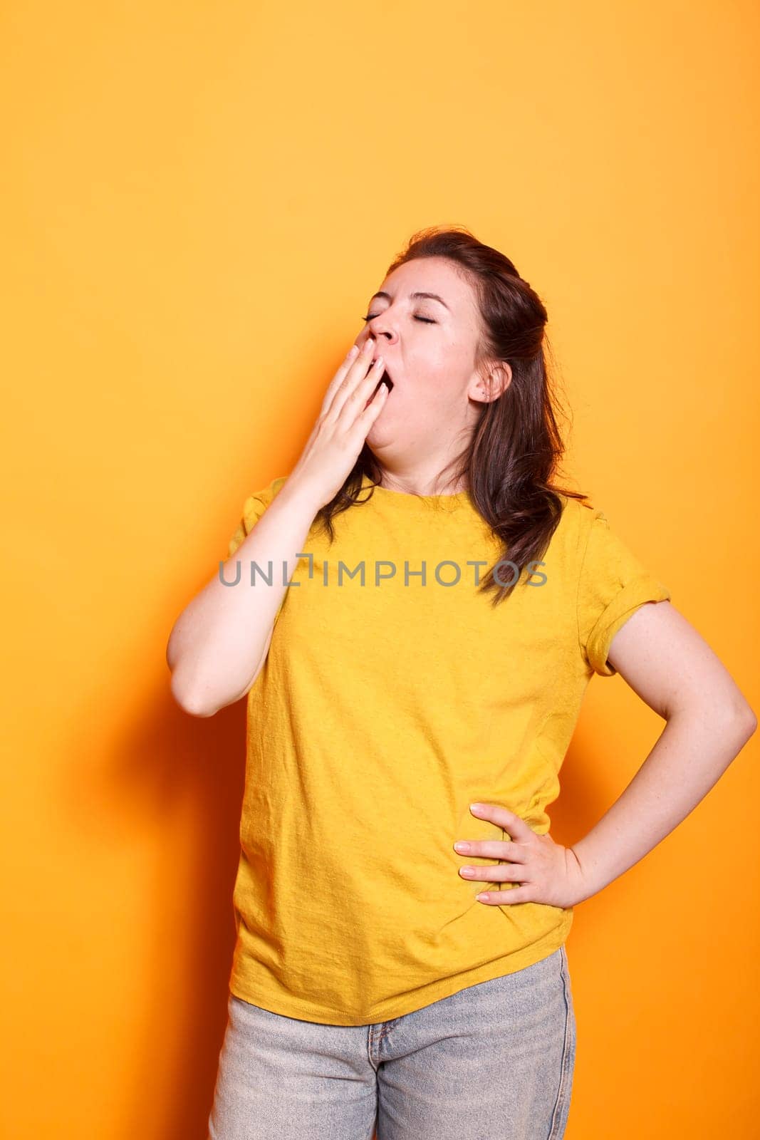 Portrait of a drowsy, young Caucasian lady in a studio, covering her mouth and yawning. Weary woman with hand on her waist, posing in front of camera and drifting off to sleep.