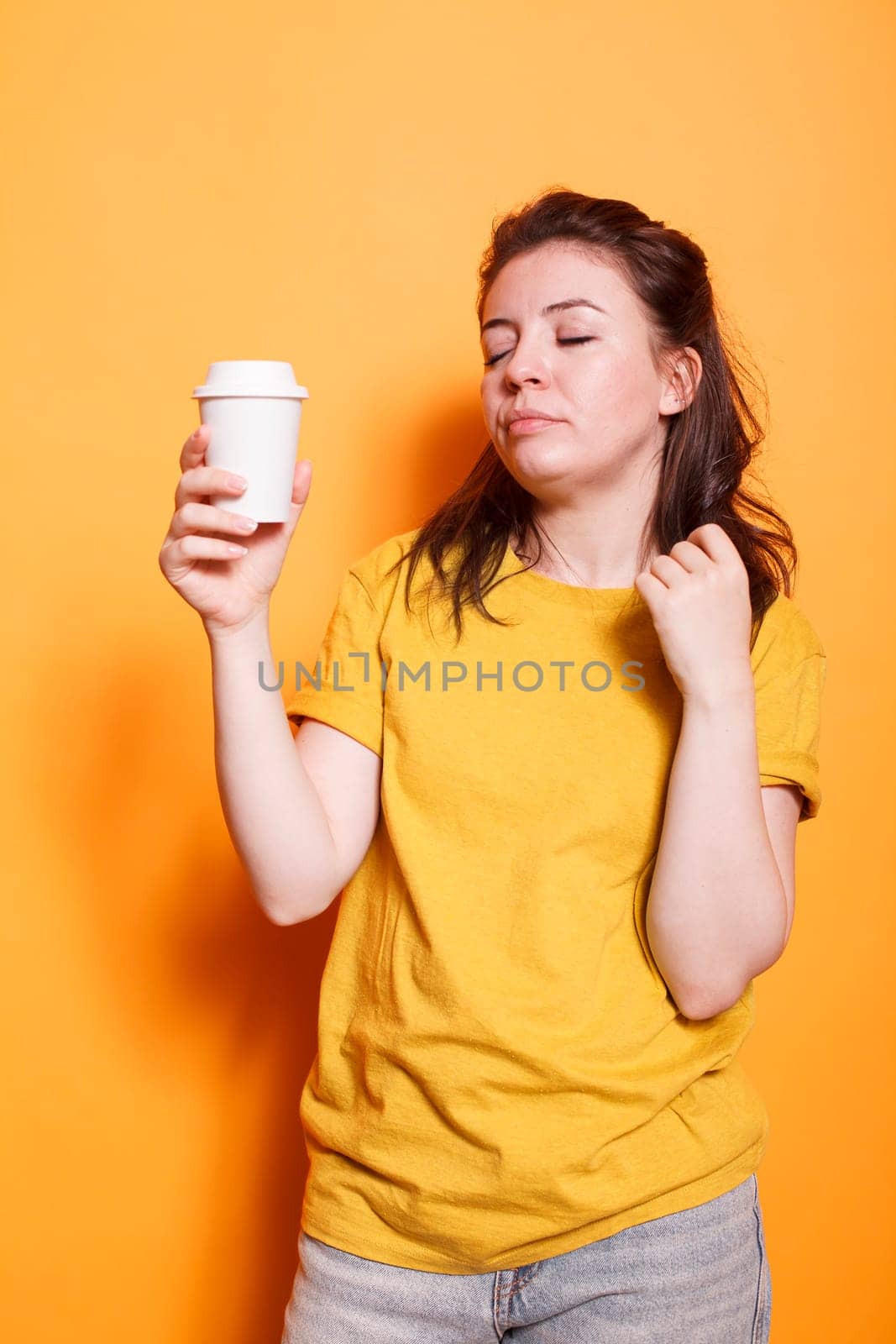 Tired woman posing against isolated orange background in casual clothes, holding coffee cup, relaxing with closed eyes. Sleepy expression of young brunette lady depicts exhaustion and weariness.