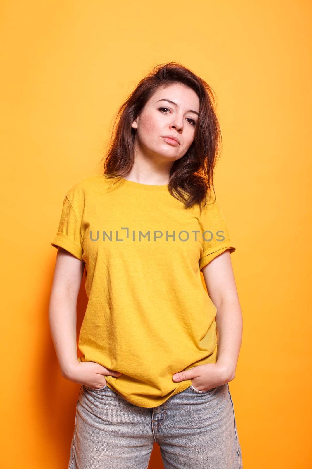 Confident woman with hands in pocket, brown hair, and casual clothing stands against an isolated orange background. Her expressive eyes and friendly demeanor are captivating.
