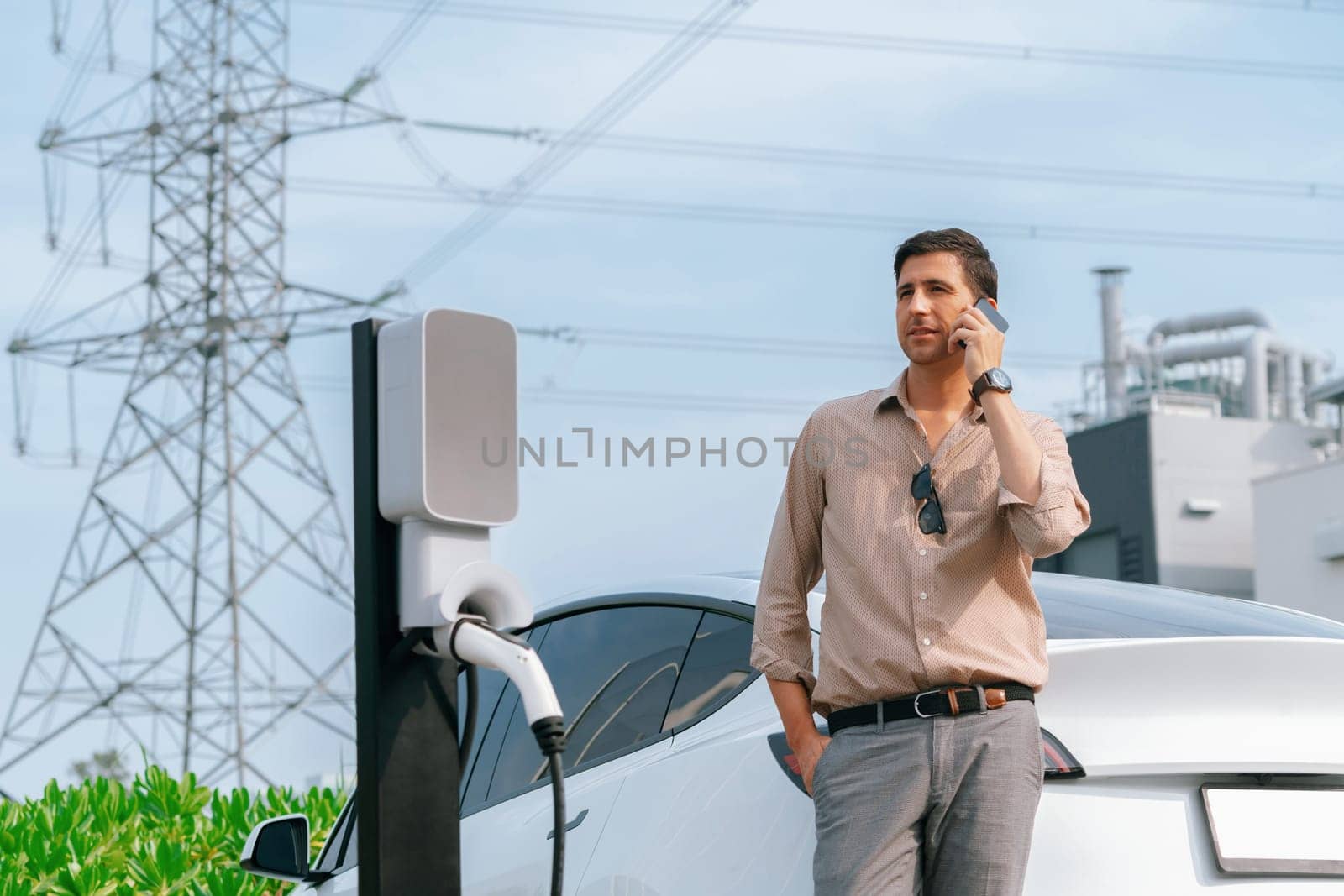 Man talking on the phone while recharge EV car battery at charging station connected to power grid tower electrical as electrical industry for eco friendly car utilization.Expedient