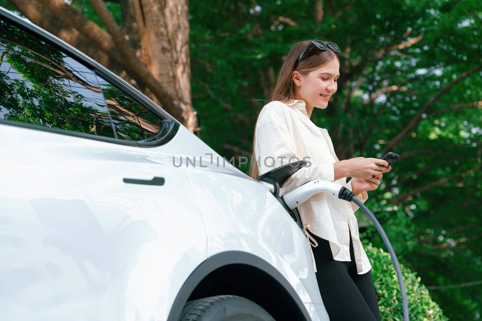 Young woman use smartphone to pay for electricity at public EV car charging station green city park. Modern environmental and sustainable urban lifestyle with EV vehicle. Expedient