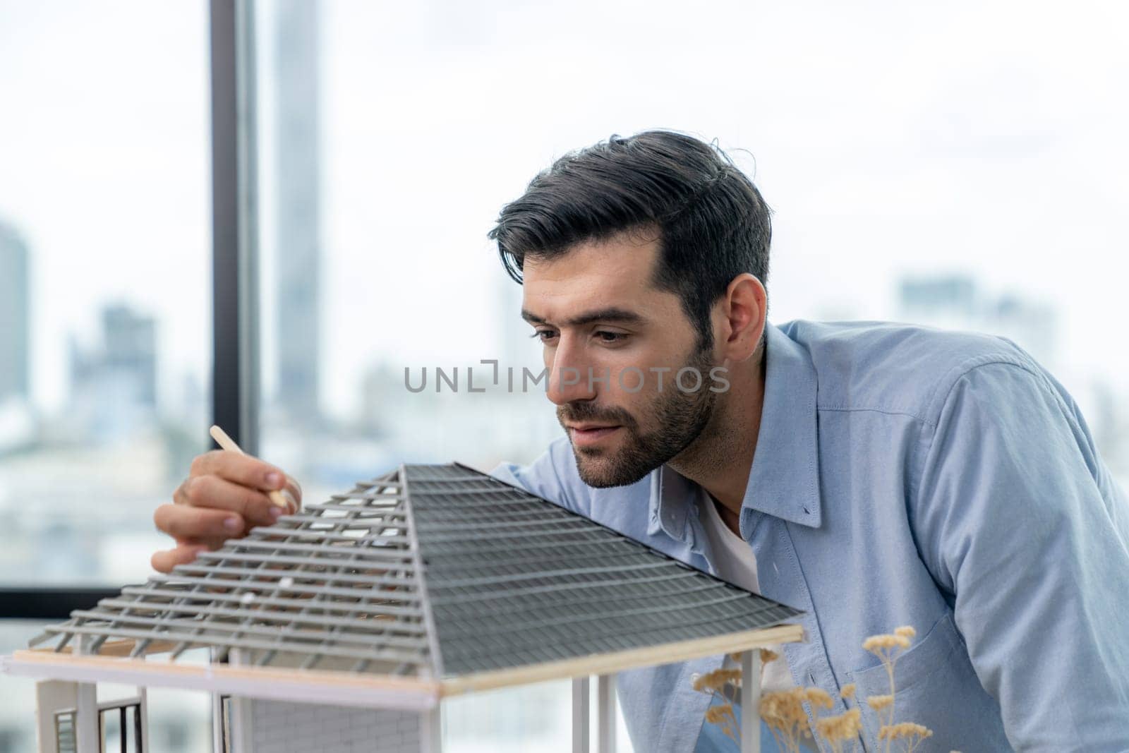 Attractive smart civil engineer measure house model roof construction. Professional architect in casual outfit looking at house model while planing building structure. Skyscraper view. Tracery.