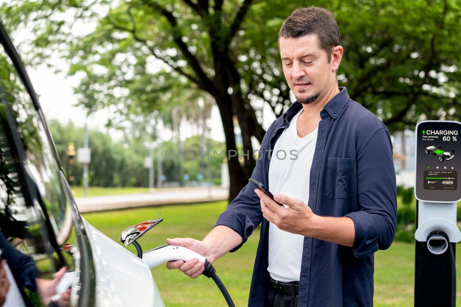 Man using smartphone online banking application to pay for electric car battery charging from EV charging station during vacation holiday road trip at national park or summer forest. Exalt