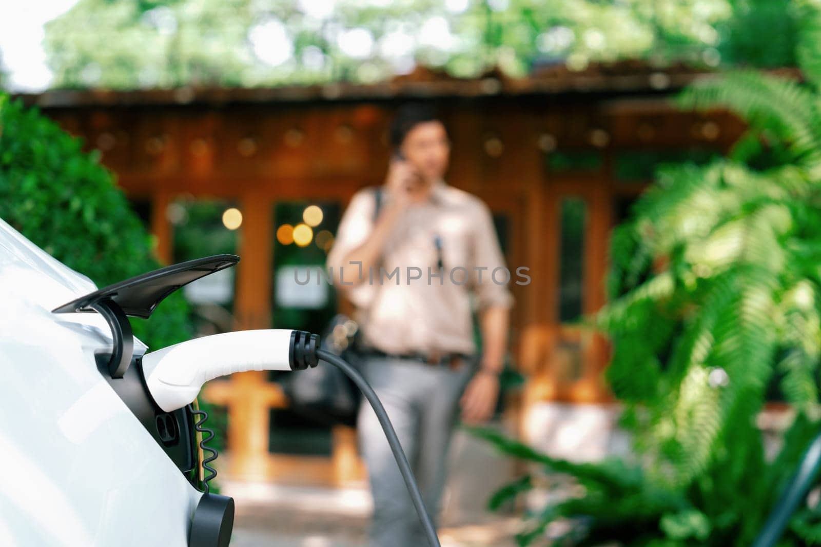 Focused EV electric car recharging at outdoor coffee cafe in springtime garden with blur background of eco friendly man, green city sustainability and environmental friendly EV vehicle. Expedient