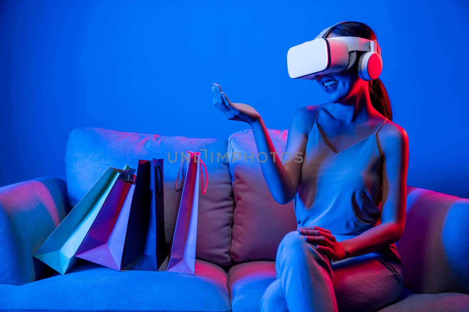 Smart female sit on sofa beside bags wear VR headset connecting metaverse, future cyberspace community technology. Elegant woman enjoy shopping products from online store in meta world. Hallucination.