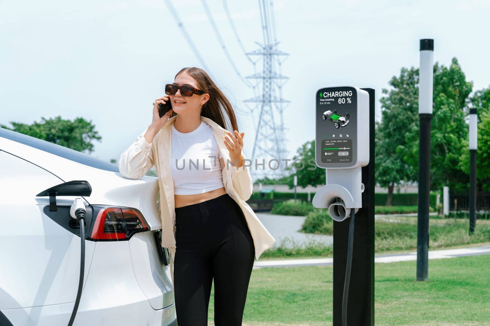 Young woman recharging EV car battery while talk on phone at charging station connected to electrical power grid tower facility as electrical industry for eco friendly vehicle utilization. Expedient