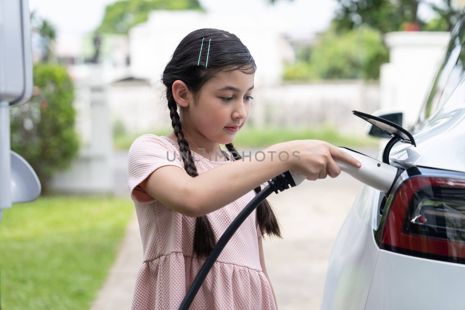 Happy little young girl learn about eco-friendly and energy sustainability as she recharge electric vehicle from home EV charging station. EV car and sustainable future generation concept. Synchronos